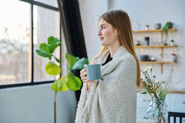 Young beautiful smiling calm woman with blanket mug, looking out winter window