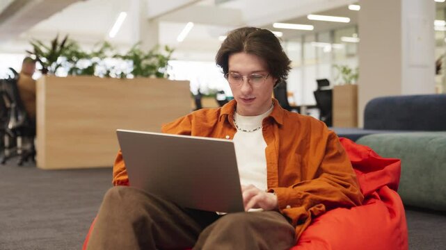 Focused Caucasian male with long hair sitting in bean chair. Typing with both hands on laptop keyboard. Showing focus in his work. Thinking about best reply to important email. Working in office.