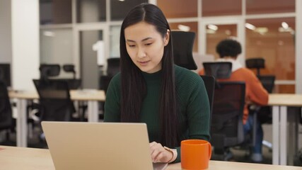 Focused Asian woman sitting in modern office. Typing on keyboard while working on laptop. Concentrating on task with serious expression. Completing assignment efficiently with full attention.