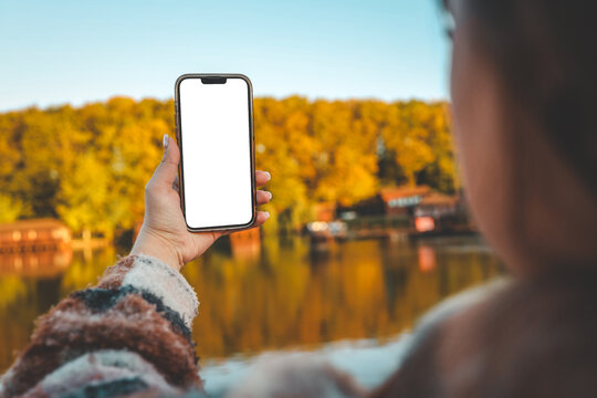 Woman holding smartphone with blank white screen outdoors near lake and autumn forest. Concept of mockup, travel and social media content creation