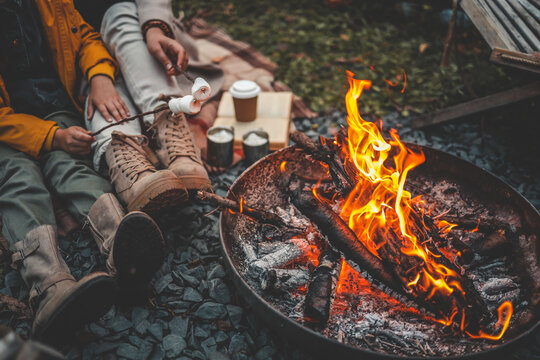 Family or friends relaxing by a campfire, roasting marshmallows on sticks. Boots, cozy clothes, hot drinks, and warm flames create a peaceful outdoor autumn evening vibe