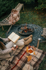 Autumn outdoor moment with a woman relaxing by the campfire, holding a book and hot drink with...