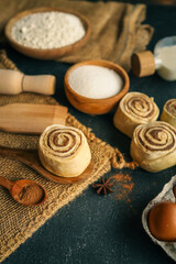 Close-up of raw cinnamon rolls on wooden spoons with baking ingredients like eggs, sugar, milk, flour, and cinnamon on rustic dark background. Homemade pastry preparation