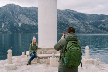 Man shoots with a smartphone a woman sitting on the step of a lighthouse on the pier. Back view