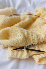 Yellow cotton knitting with knitting needles on white sheet. Soft yellow knitwear with circle needles. Knitting pattern background. Knitting lifestyle. Cozy hobby. Warm knitwear. Craft product concept