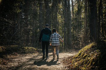 Obraz premium A man and woman walk side by side, holding hands on a sunlit forest trail. They are warmly dressed, enjoying a peaceful moment in nature.