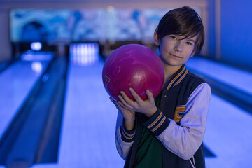 A young boy stands confidently in a bowling alley, holding a bright pink bowling ball under neon lighting, ready for his turn on the lanes.