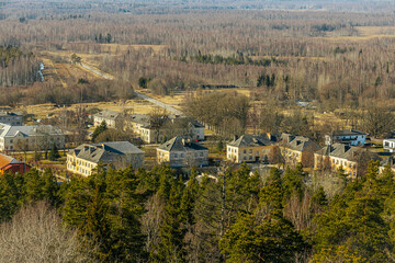 Obraz premium Elevated view of a quiet Zilaiskalns town with modest residential buildings, surrounded by dense woodland and leafless trees in early spring, under clear skies.