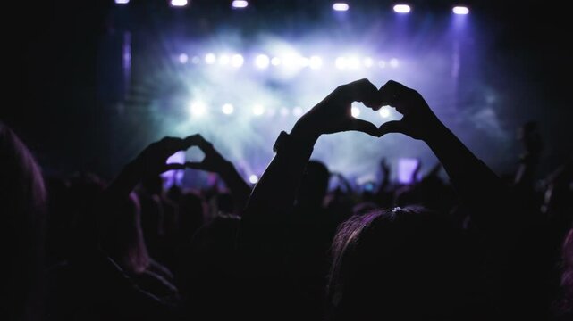 Concerthall show. Dark silhouette of female hands in the shape of heart fingers, blue backlight from the stage. Medium close-up shot. 