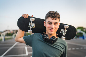 One man young adult caucasian teenager stand outdoor with skateboard on his shoulder and headphones...