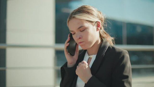 Business lady on phone displays frustration during conversation, puffing cheeks and raising hand in anger while walking past mall exterior with glass facade