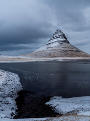 Kirkjufell mountain mirrored in a partially frozen lake under dramatic winter skies, Snæfellsnes Peninsula, Iceland. Iconic and moody Nordic scenery