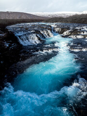 Bruarafoss Waterfall, Iceland: Turquoise glacial waters flow over rugged rocks, creating a mesmerizing cascade. Surrounded by winter landscape, it’s a natural wonder