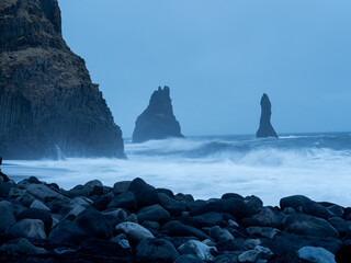 Reynisfjara Beach at dusk, with the iconic Reynisdrangar sea stacks standing against turbulent waves, framed by dark basalt columns and scattered volcanic rocks