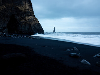 Reynisfjara Beach at dusk, featuring dramatic black sand, basalt cave, rugged cliffs, and the iconic Reynisdrangar sea stacks emerging from the turbulent Atlantic Ocean