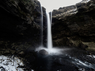 Kvernufoss waterfall plunges into a dark, rocky basin surrounded by rugged basalt cliffs. The flowing water contrasts with the snow-dusted landscape, highlighting Iceland’s raw beauty