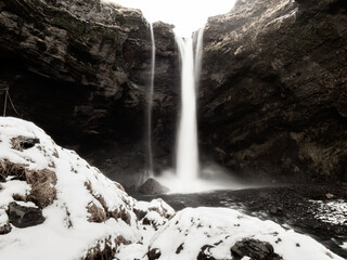 Kvernufoss waterfall plunging from a rocky cliff into a misty pool, framed by snow-covered boulders. A hidden winter wonder in Iceland's wild landscape