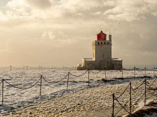 Golden light bathes Dyrhólaey lighthouse, Iceland. The iconic red-topped structure stands against a snowy, frozen landscape, exuding solitude and tranquility in winter