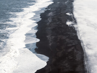 Aerial view of the black sand beach at Dyrhólaey, Iceland. Waves crash against the dark shoreline, blending with snow-covered sand and leaving visible tire tracks