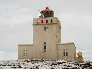 Historic Dyrhólaey lighthouse in Iceland, perched on a snow-dusted hill. The iconic red dome contrasts with the muted winter landscape, symbolizing resilience against the elements