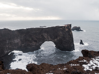Dyrhólaey Arch in Iceland, a dramatic natural rock arch stretching over the Atlantic Ocean, surrounded by rugged cliffs, crashing waves, and snow-covered landscape.