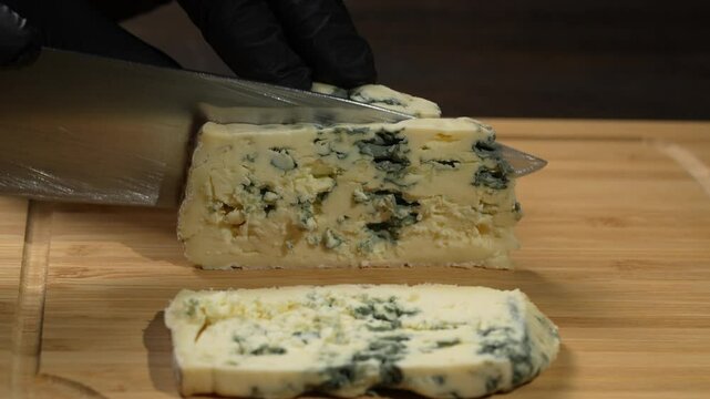 The cook cutting the blue cheese dorblu into slices with knife on cutting wooden board, close up. Delicious blue cheese