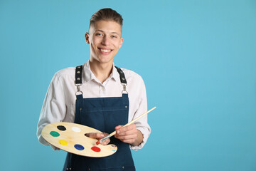 Smiling man with wooden palette and paintbrush on light blue background. Space for text