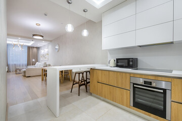 Stylish interior design of the studio apartment. White table with high chairs, kitchen set. A wide passage into the living room.
