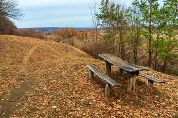 Fototapeta premium Old wooden table with benches near a hiking trail on a hill near a forest. Picturesque countryside landscape in Ukraine. Fallen autumn leaves on yellow dry grass