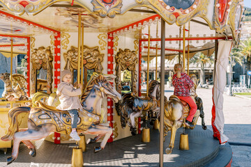 Little girls ride on a colorful carousel holding on to poles. Side view