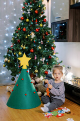 Little girl holds a felt toy in her hands while sitting on the floor near a toy Christmas tree