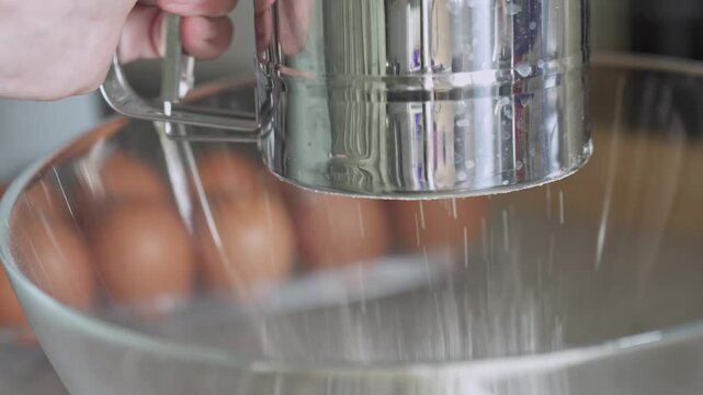 Flour being sifted into clear glass bowl with a metal sifter during cooking