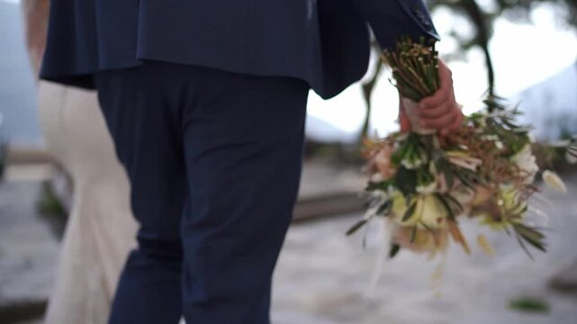 Bouquet of flowers in the hand of the groom walking with the bride along the road. Back view