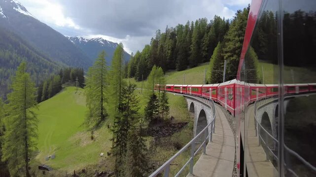 Grisons, Switzerland - April 12. 2024: Bernina Express, red train pass over a old stones bridge with arches in beautiful Swiss Alps landscape