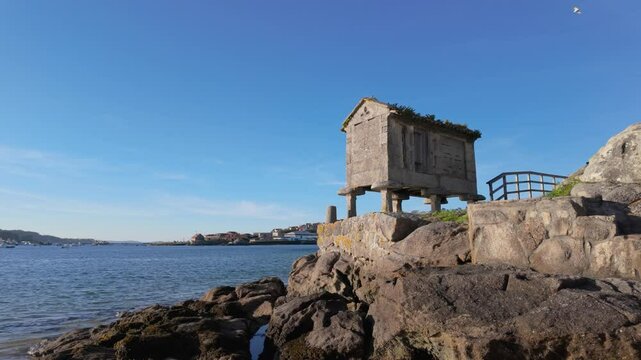 Picturesque horreo, a traditional galician raised granary, stands on the shores of Ald&aacute;n, showcasing its unique architectural style against a serene backdrop of water and blue sky