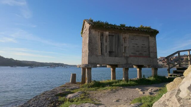 Picturesque horreo, a traditional galician raised granary, stands on the shores of Ald&aacute;n, showcasing its unique architectural style against a serene backdrop of water and blue sky
