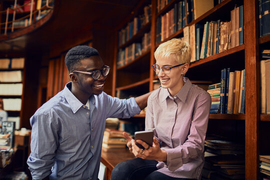 Diverse student friends using smartphone and laughing together in library