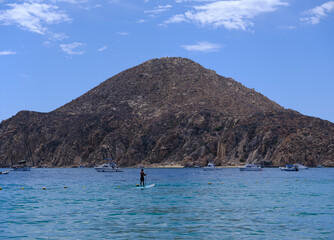 Fototapeta premium A serene coastal scene with a person paddleboarding in calm water, surrounded by boats and overlooked by a large rocky mountain under a bright blue sky.