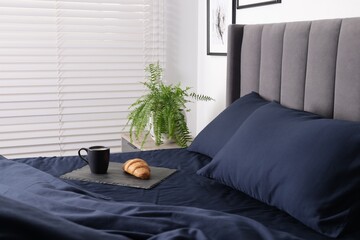 Tasty coffee and croissant on bed with dark blue silk linens indoors