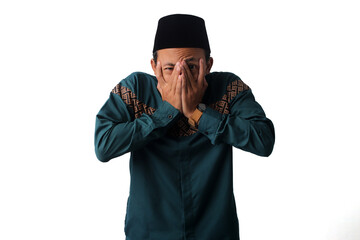 Asian Muslim man covers his face, expressing fear and concern about actions that could break his...