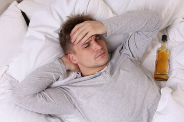 Unhappy young man with bottle of whiskey suffering from hangover on bed at home, top view