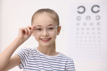 Little girl wearing glasses near vision test chart at ophthalmologist office, space for text