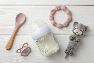 Feeding bottle with milk, needle felted cat, pacifier, teether and spoon on white wooden table,...
