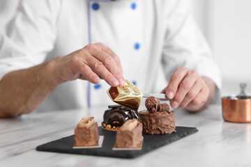 Elderly pastry chef decorating tasty desserts with edible gold leaf sheet at table in kitchen, closeup