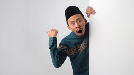 Surprised Asian Muslim man peeking from behind a white wall, pointing with his thumb to the side while showing an amazed expression with his mouth open. Isolated on a white background