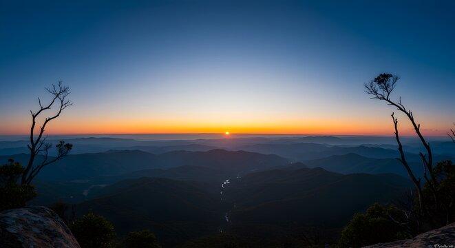 Panoramic sunrise view over mountain range, silhouetted trees, gradient sky
