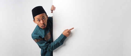 A surprised Asian Muslim man peeking from behind a white wall while pointing at the empty copy space, as if recommending something. Isolated on a white background