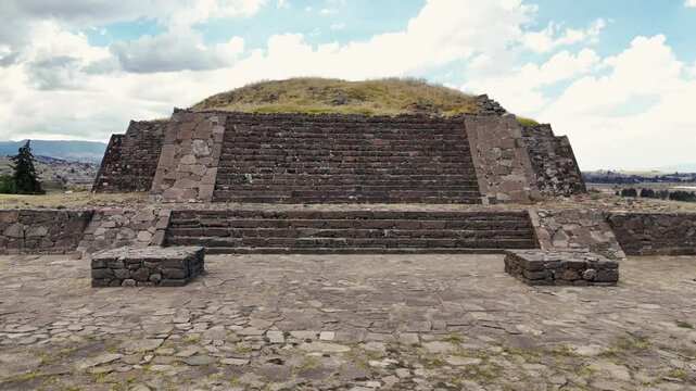 Pyramids of Tlaloc with village and mountains in the background at the archaeological site of Calixtlahuaca in Mexico. Drone camera approaches pyramid at low altitude and rises revealing the village
