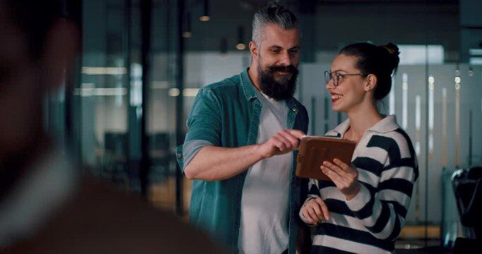 Male senior coach or speaker sharing business ideas and collaborating with a diverse team in a modern office, presenting a project on a tablet during a brainstorming session in a startup hallway