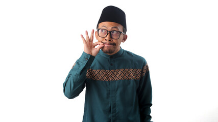 Funny Asian Muslim man making a gesture with his hand as if zipping his lips, playfully promising to keep a secret while looking away at empty copy space, isolated on a white background